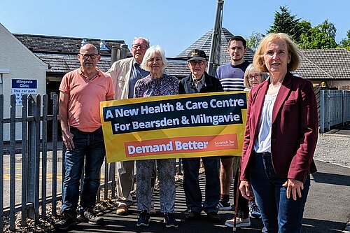 Susan Murray in front of a group of campaginers who are holding up a banner that reads "A new health centre for Bearsden and Milngavie. Demand Better. Scottish Liberal Democrats".